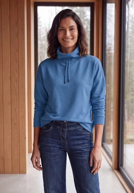 Langarm Shirt mit Rollkragen und Tunnelzug north blue Eine Frau im blauen Pullover und Jeans steht vor großen Fenstern in einem Holzraum und lächelt.