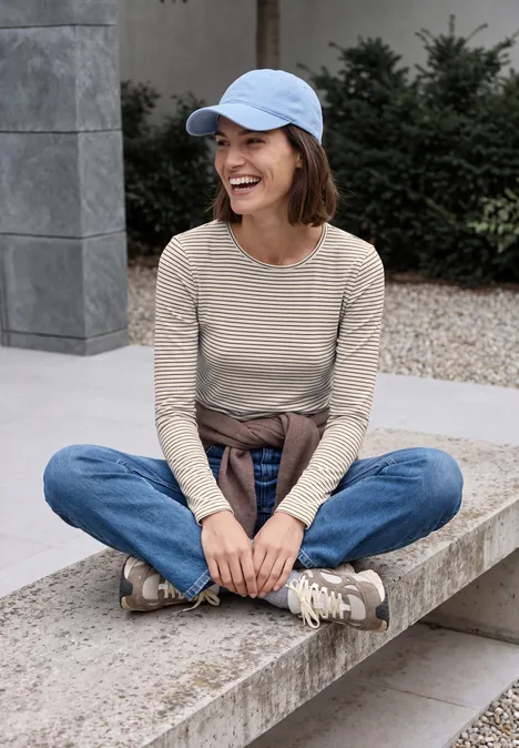 T-shirt à manches longues, col rond et rayures pearl beige Lachende Frau sitzt im Schneidersitz auf einer Steinbank, trägt Jeans, beiges Langarmshirt und Cap.
