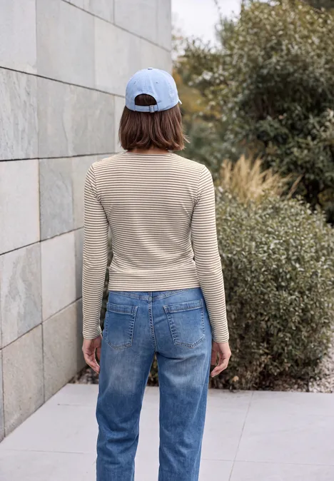 T-shirt à manches longues, col rond et rayures pearl beige Frau steht mit dem Rücken zur Kamera, trägt Jeans, Beige-Langarmshirt und Baseballcap, vor einer Mauer und Büschen.