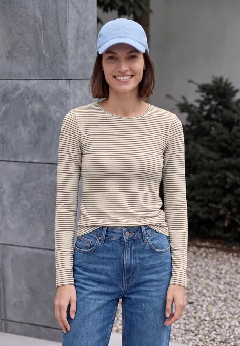 T-shirt à manches longues, col rond et rayures pearl beige Junge Frau mit weißer Kappe, beige gestreiftem Shirt und blauen Jeans vor grauer Wand.