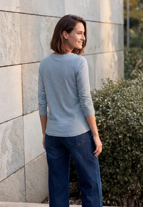 Shirt met lange mouwen, ronde hals en strepen cosmopolitan blue Frau in grauem Dreiviertelshirt und blauer Jeans lehnt an einer Steinwand und schaut seitlich.
