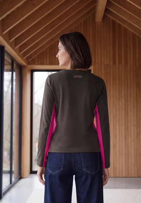 Frau steht in Holzbau, trägt olivgrünen Pullover mit pinken Seiteneinsätzen und Jeans.
