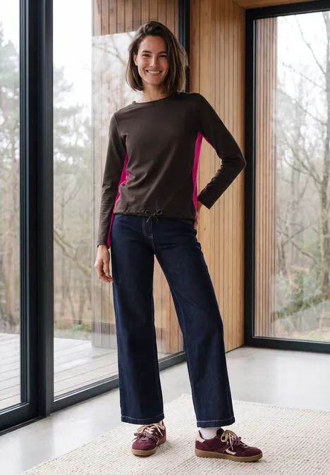 Eine lächelnde Frau in modernem Longsleeve und weiten Jeans steht vor großen Fenstern in einem hellen Raum.