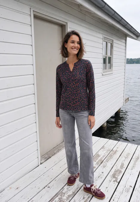Eine Frau steht auf einem Holzsteg vor einem weißen Bootshaus am See.