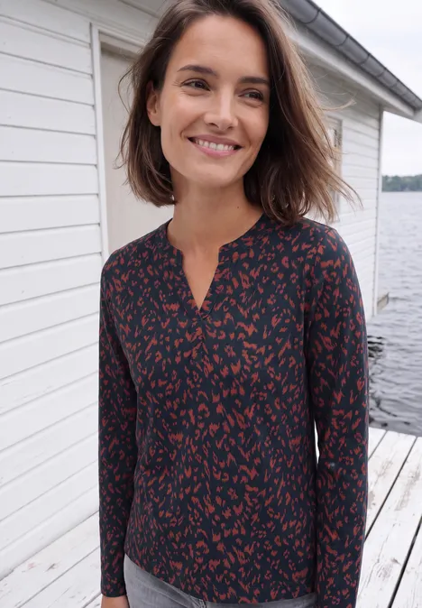 Eine lächelnde Frau mit schulterlangen braunen Haaren trägt eine dunkelblau-rote Bluse und steht auf einem Steg am Wasser.