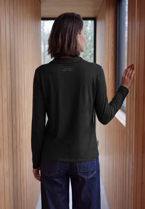 Person von hinten in schwarzem Langarmshirt und Jeans, steht im hellen Holzflur am Fenster.