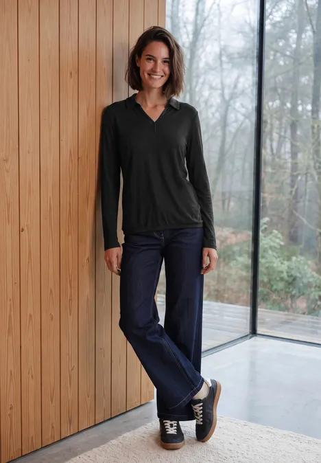 Eine Frau lehnt lächelnd in Jeans und schwarzem Langarmshirt an einer Holzwand vor großen Fenstern.