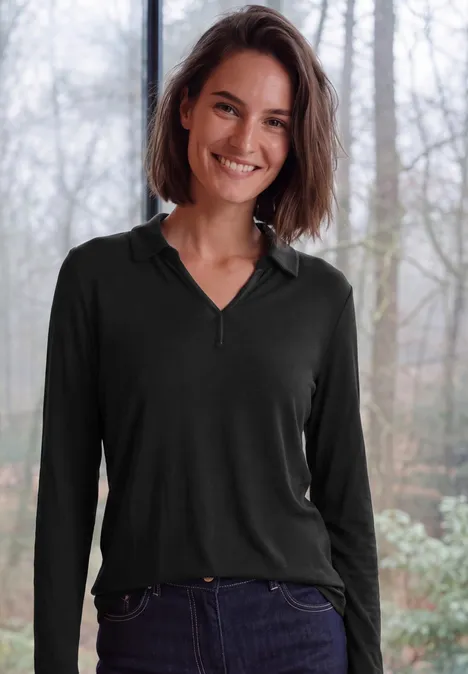 Eine lächelnde Frau in schwarzem Langarmshirt und Jeans vor unscharfem Waldhintergrund.