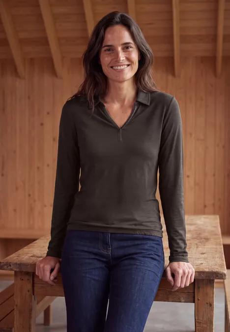 Frau lehnt lächelnd an einem Holztisch vor einer hölzernen Wand, trägt langärmeliges Oberteil und Jeans.