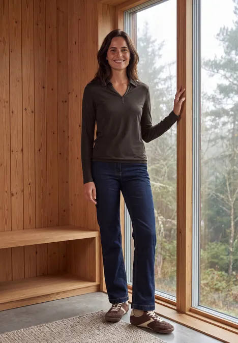 Frau in lässiger Kleidung (Pullover, Jeans, braune Schuhe) steht in modernem Holzinnenraum am Fenster.