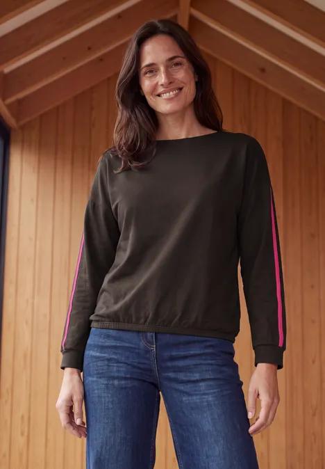 Eine lächelnde Frau in Jeans und langärmeligem Oberteil steht vor einer Holzwand.