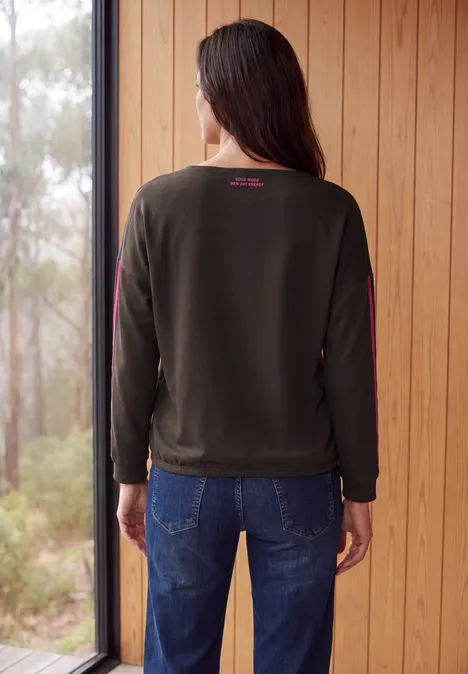 Frau von hinten in dunklem Sweatshirt und Jeans, steht vor Holzwand und Blick auf bewaldete Umgebung.