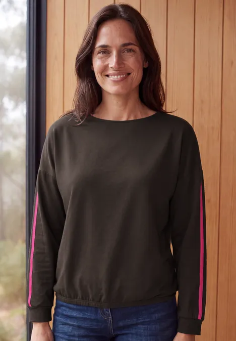 Eine lächelnde Frau in grünem Sweatshirt und Jeans vor einer Holzwand.