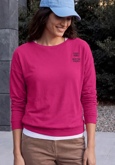 Jonge vrouw draagt roze trui, wit shirt, beige broek en blauwe pet, glimlacht buiten voor een stenen muur.