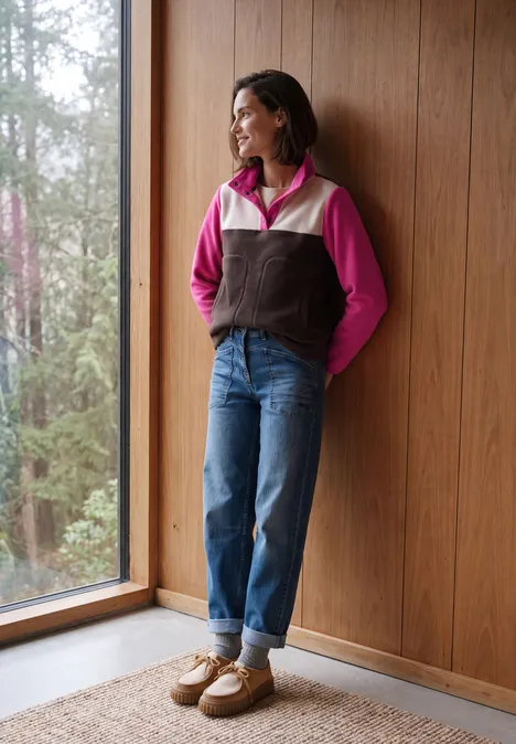 Frau in Jeans, pink-brauner Jacke und Hausschuhen steht lächelnd am Fenster vor Holzpaneelen.
