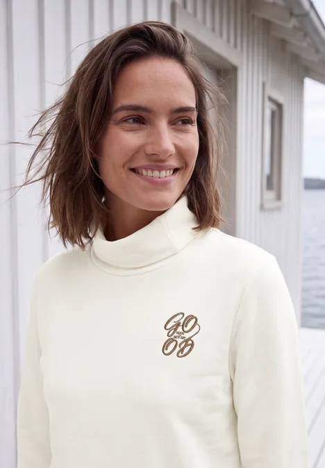 Vrouw in lichte coltrui met stijlvol logo, lachend voor een wit houten huisje aan het water.