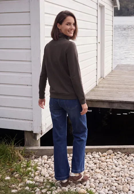 Een glimlachende vrouw in spijkerbroek en donkere trui staat op een steiger naast een wit bootshuis.