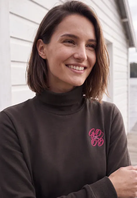 Een glimlachende vrouw met donkerbruin haar staat voor een wit huis en draagt een donkere coltrui met een roze logo.