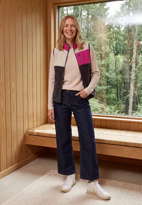 Frau in Holzinterieur neben Panoramafenster, trägt Farbblock-Weste, Pullover, weite Jeans und weiße Schuhe.