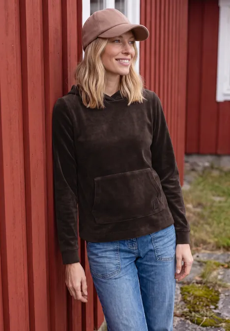 Een vrouw in een donkerbruine hoodie, spijkerbroek en pet leunt glimlachend tegen een rode houten muur.