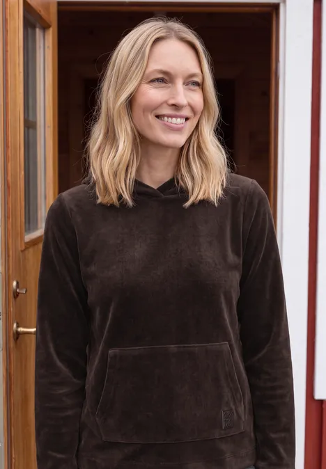 Een lachende vrouw met blond haar draagt een bruin sweatshirt en staat voor een open houten deur.