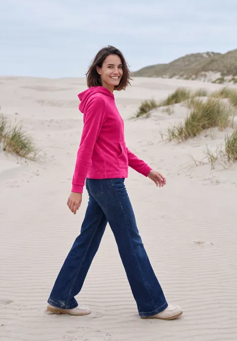 Kobieta w różowej bluzie z kapturem i jeansach spaceruje po plaży.