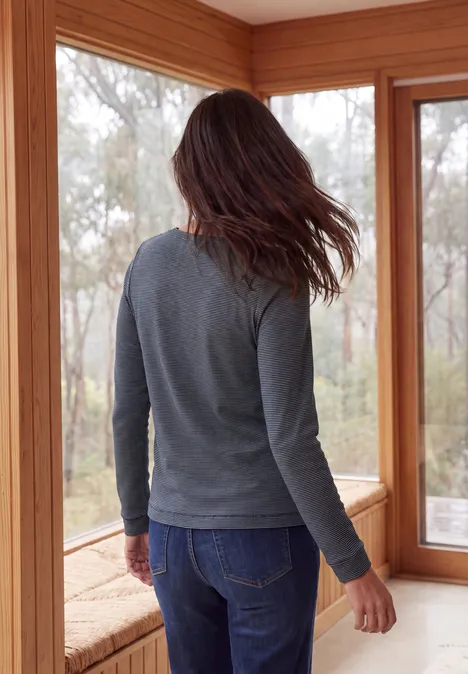 Eine Person steht mit dem Rücken zur Kamera an einer Fensterbank, trägt grauen Pullover und Jeans.