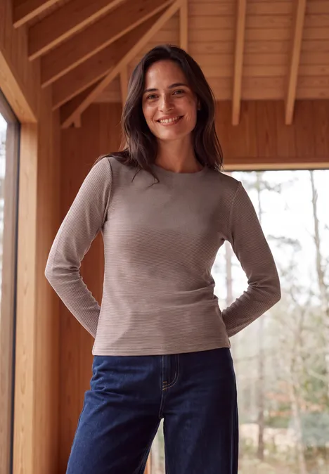 Eine lächelnde Frau in grauem Shirt und Jeans steht vor einer Holzwand mit Blick ins Freie.
