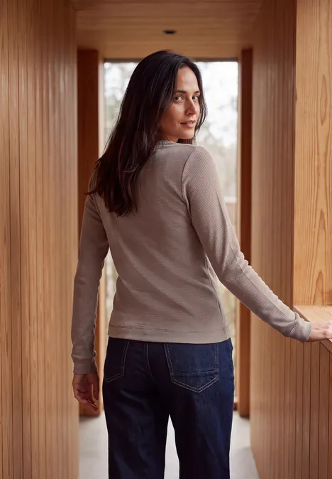 Eine Frau im hellen Langarmshirt und Jeans steht in einem hölzernen Gang und blickt über die Schulter.