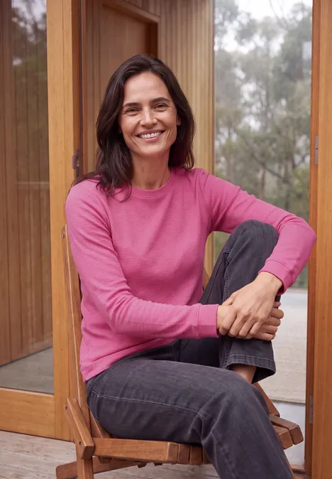 Eine lächelnde Frau in einem pinken Pullover sitzt entspannt auf einem Holzstuhl vor großen Fenstern.