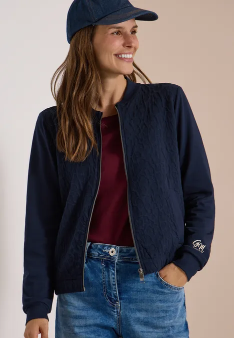 Blouson mit Strukturmix und Elastiksaum urban dark blue Junge Frau mit dunkelblauer Steppjacke, rotem Shirt, Jeans und Cap, lächelnd zur Seite blickend.