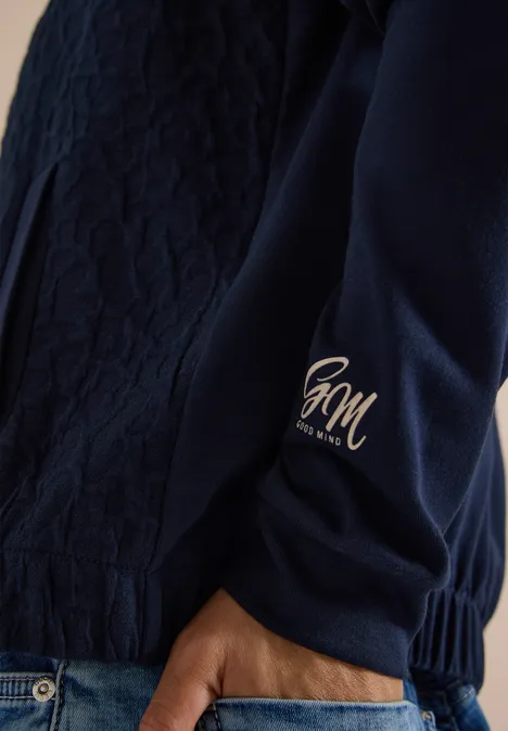 Blouson mit Strukturmix und Elastiksaum urban dark blue Nahaufnahme eines dunkelblauen Steppjackenärmels mit goldfarbenem HM-Logo am Bündchen und Hand in Jeans