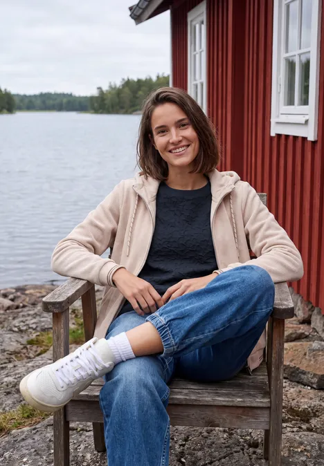 Shirt mit Boat Neck und Strukturmix urban dark blue Eine lächelnde Frau in Jeans und Kapuzenjacke sitzt auf einem Holzstuhl vor einer roten Hütte am See.