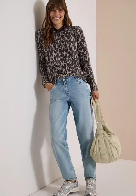 Langarm Shirt mit Fake-Kapuze truffle brown Frau in gemustertem Oberteil, blauen Jeans und Tasche, lächelnd an die Wand gelehnt.