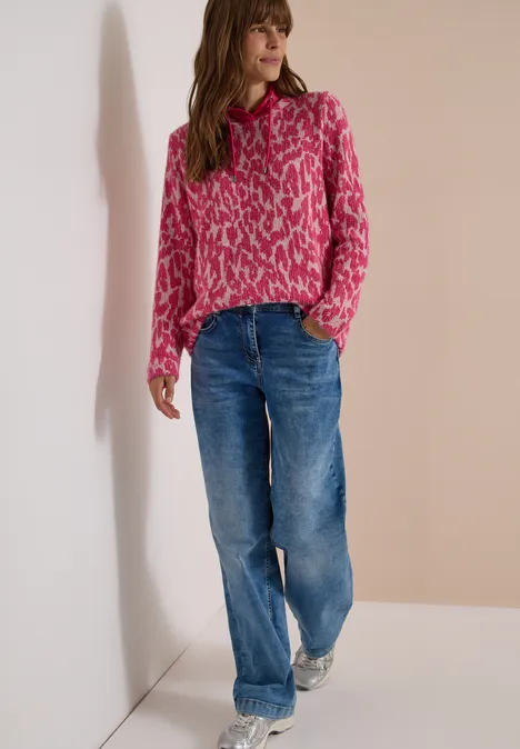 Langarm Shirt mit Fake-Kapuze pink flash Frau steht an Wand, trägt pinkgemusterten Pullover und lockere blaue Jeans.