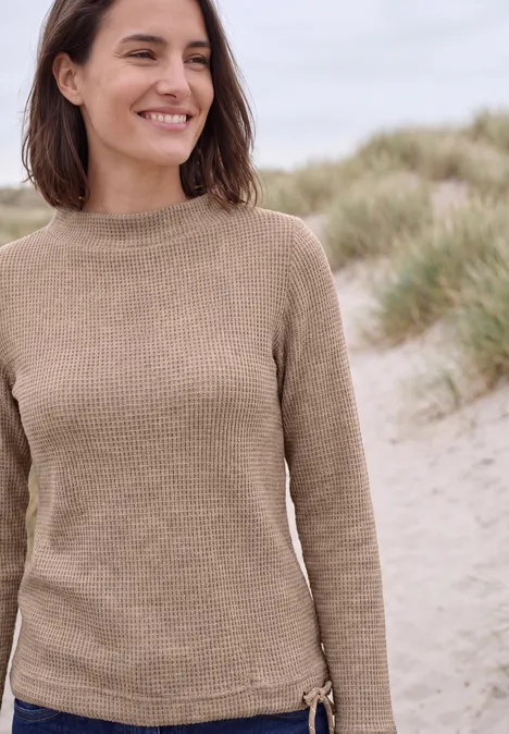 Une femme souriante en pull beige se tient devant des dunes.