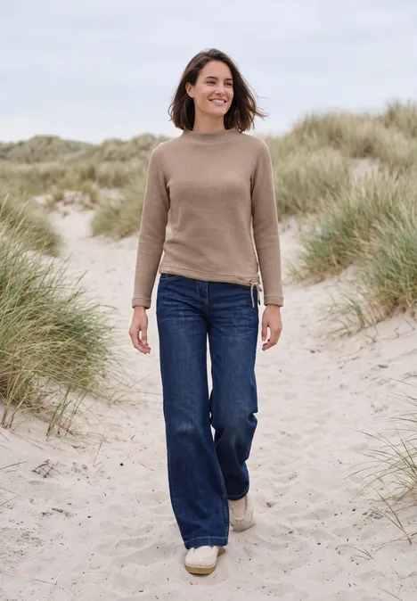 Une femme marche sur des dunes herbeuses au bord de la mer, en pull, jean et baskets blanches.