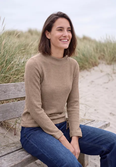 Femme assise sur un banc de plage, souriante en pull et jean, herbes et dunes en arrière-plan.