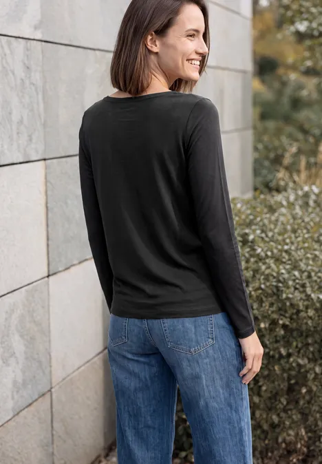 Vrouw in zwart shirt en blauwe spijkerbroek, deels van achteren gezien bij stenen muur en struiken.