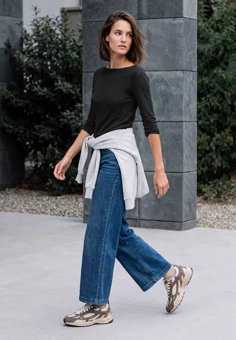 Vrouw in zwarte top en jeans met sneakers en grijze trui om de heupen, lopend voor modern gebouw.