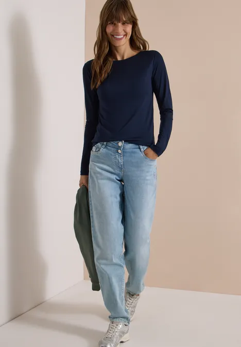 Langarm Shirt in Unifarbe mit Boat Neck urban dark blue Frau in blauem Shirt und Jeans steht lächelnd vor beigem Hintergrund, hält eine Jacke.