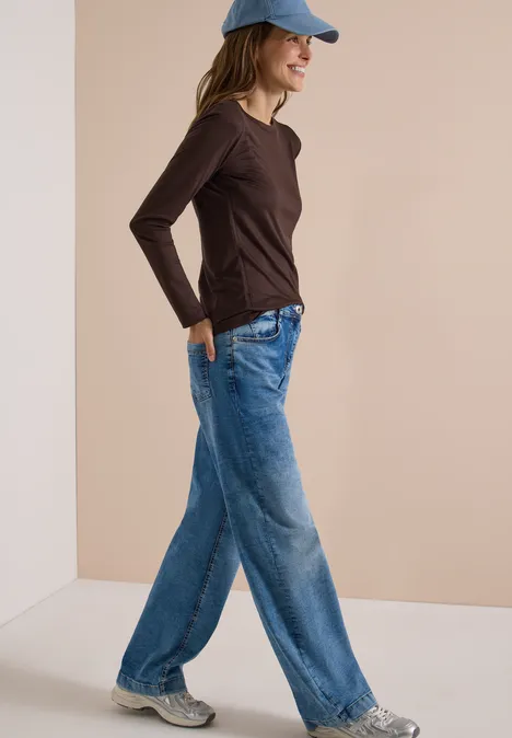 Langarm Shirt in Unifarbe mit Boat Neck truffle brown Frau in blauer Jeans, braunem Langarmshirt und Cap, die lächelnd vor hellem Hintergrund geht.