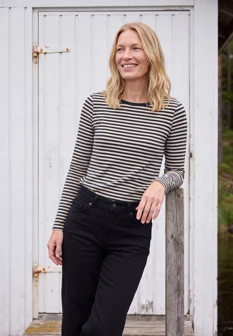 Langarm Shirt mit Boat Neck und Streifen urban dark blue Eine lächelnde blonde Frau in gestreiftem Langarmshirt und schwarzer Hose lehnt entspannt an einer weißen Holztür.