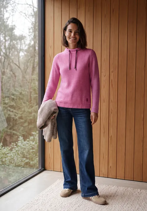 Shirt in Rippstruktur mit Funnel Neck pink flash melange Frau im pinken Kapuzenpullover mit weitem Jeans und Hausschuhen steht vor Holzvertäfelung neben großem Fenster.