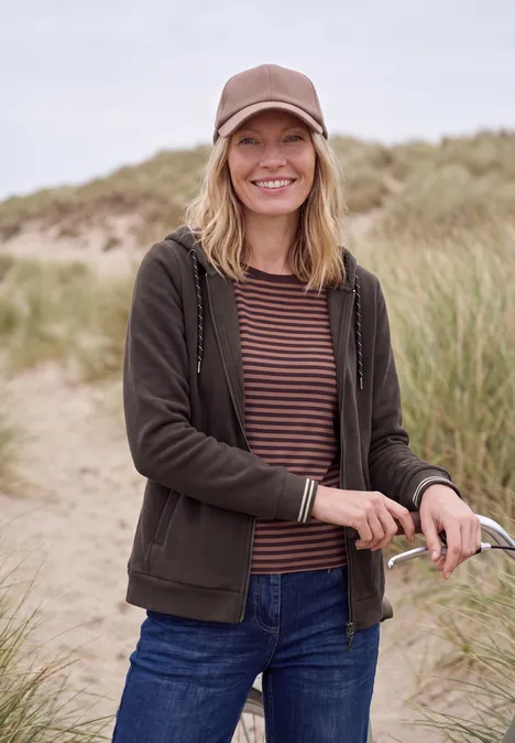 Une femme souriante en casquette et veste dans les dunes, tenant ses lunettes de soleil.