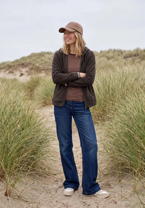 Une femme en jean, cardigan et casquette sourit les bras croisés au milieu de dunes herbeuses.