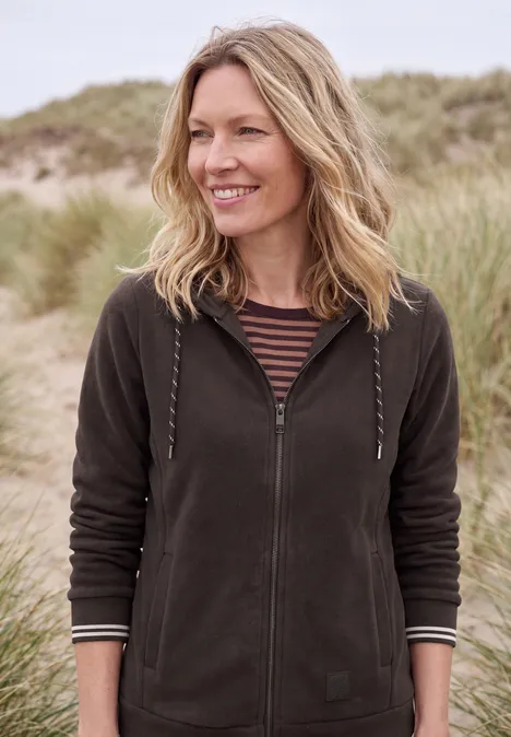 Une femme souriante en veste à capuche se tient devant des dunes de sable et des herbes de plage.