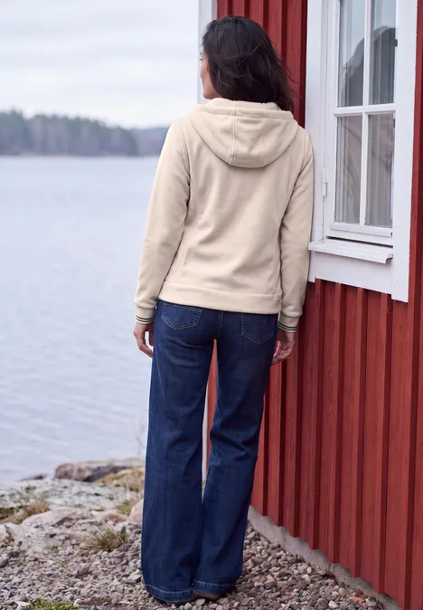 Langarm Fleece Jacke mit Kapuze cashmere beige Person mit beiger Kapuzenjacke und blauer Jeans steht mit Blick auf einen See neben rotem Holzhäuschen.