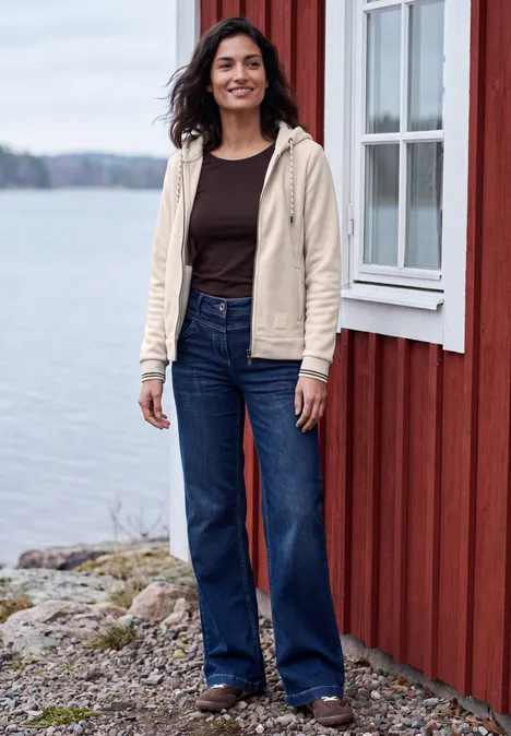 Langarm Fleece Jacke mit Kapuze cashmere beige Frau in beiger Kapuzenjacke und weiten Jeans steht lächelnd vor einem roten Haus am Wasser.