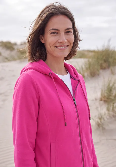 Langarm Fleece Jacke mit Kapuze pink flash Frau steht lächelnd am Strand in rosa Kapuzenjacke und weißem Shirt, Dünen im Hintergrund.